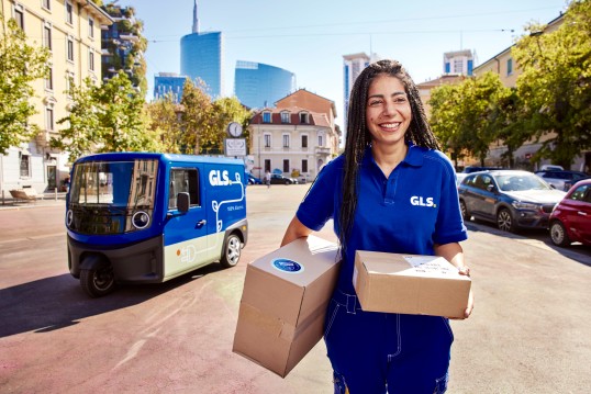 Livreuse GLS souriante portant des colis devant un véhicule de livraison GLS en ville