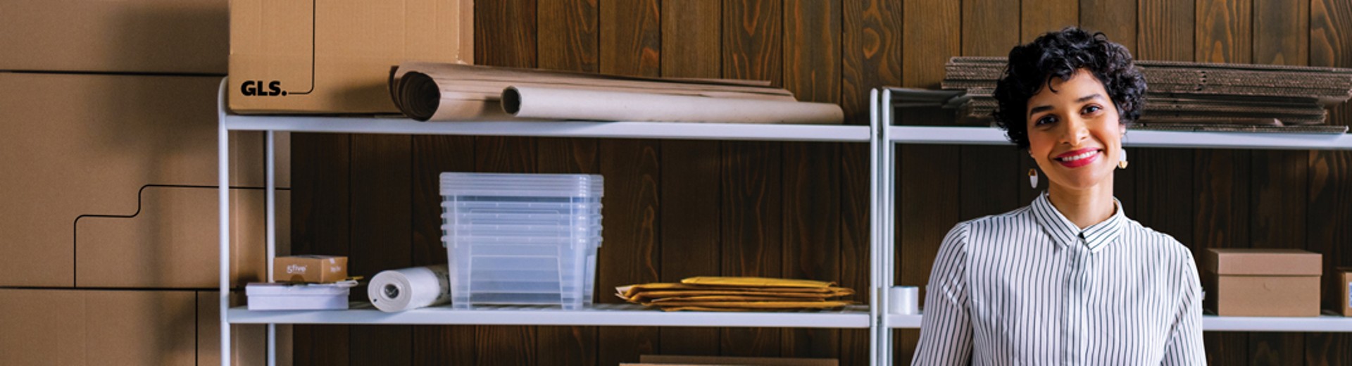 A woman standing at a table in a small warehouse, holding a cardboard shipping box. The workspace is filled with shelves of packing materials, stacked boxes, and shipping supplies.
